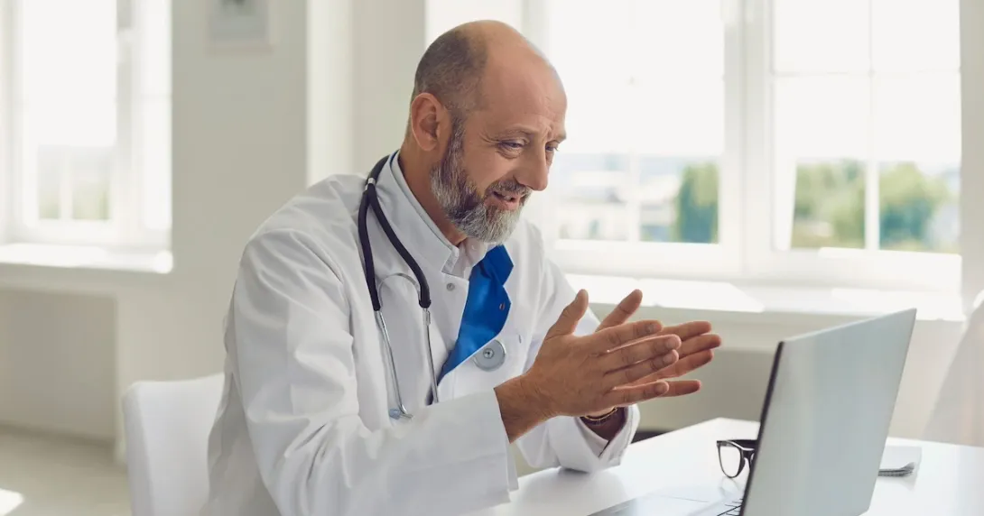 Healthcare provider sitting at a desk while wearing a lab coat and a stethoscope around their neck and speaking with someone on a computer